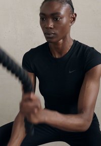 Črna prilagojena športna majica Nike s kratkimi rokavi. Posameznik se ukvarja s telovadbo in drži vrv. Gladka tekstura, preprost dizajn.