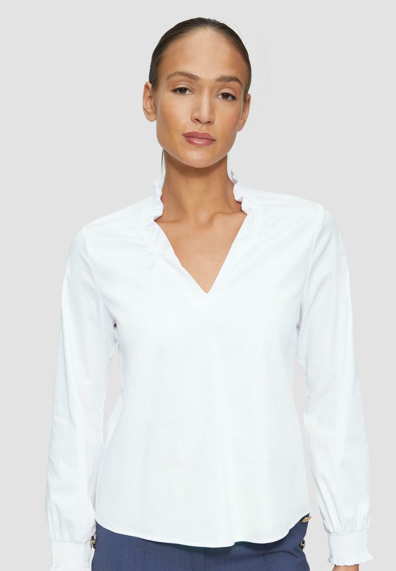 Weiße Bluse mit langen Ärmeln, einem Rüschenausschnitt und V-Ausschnitt. Weicher, glatter Stoff mit lockerer Passform und elastischen Bündchen.