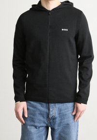 Schwarze Zip-Hoodie aus weichem Stoff, mit geripptem Textur an den Schultern, kleinem weißen Logo und kontrastierenden Bündchen.