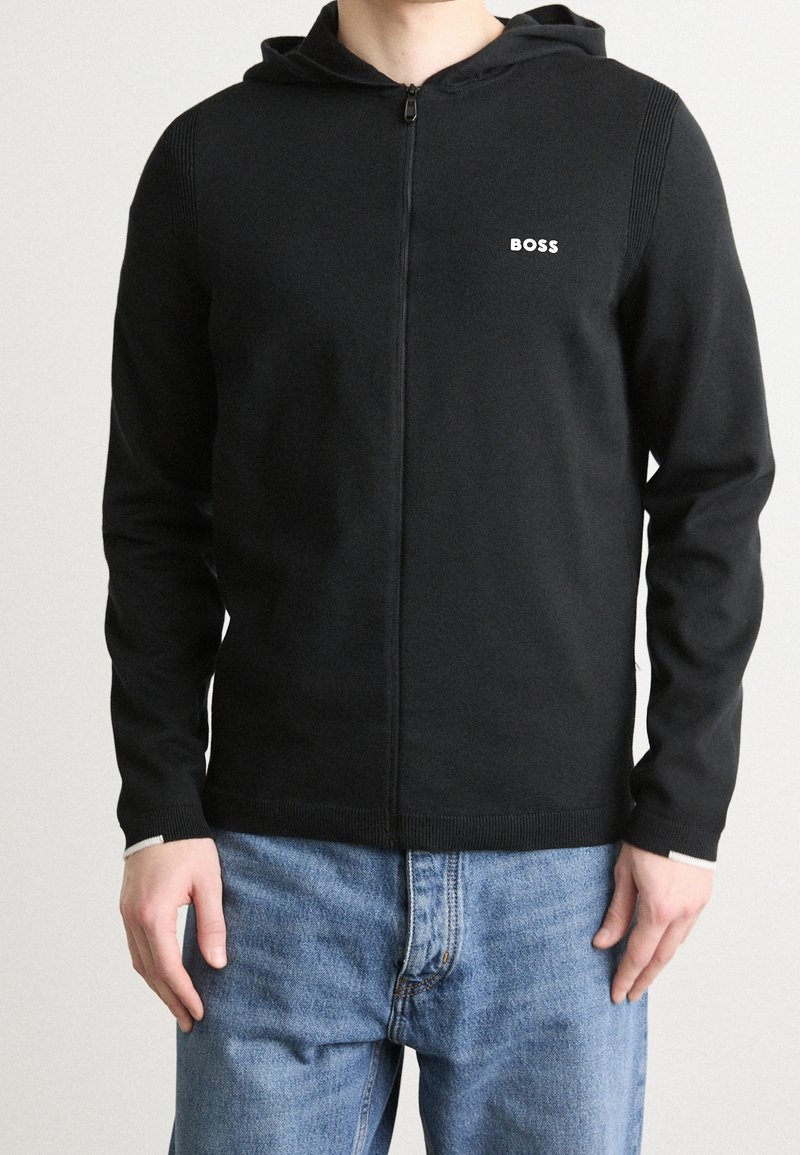 Schwarze Zip-Hoodie aus weichem Stoff, mit geripptem Textur an den Schultern, kleinem weißen Logo und kontrastierenden Bündchen.