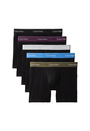 Muskuløs mand står barfodet iført sorte Calvin Klein boxershorts mod en ensfarvet grå baggrund.