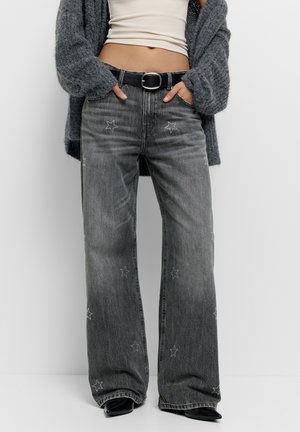 Femme portant un jean gris foncé à jambes larges avec des motifs étoiles, ceinture noire, crop top blanc et cardigan gris, mains dans les poches.