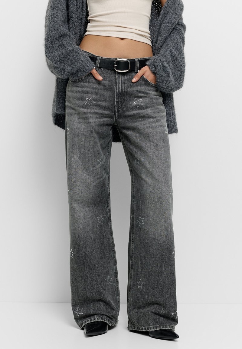 Femme portant un jean gris foncé à jambes larges avec des motifs étoiles, ceinture noire, crop top blanc et cardigan gris, mains dans les poches.