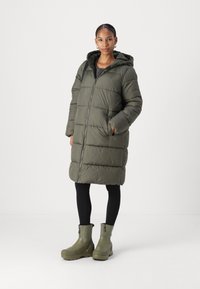 Cappotto verde stile puffer con cappuccio, zip frontale e tasche laterali. Abbinato a leggings neri e stivaletti verdi. Texture liscia.