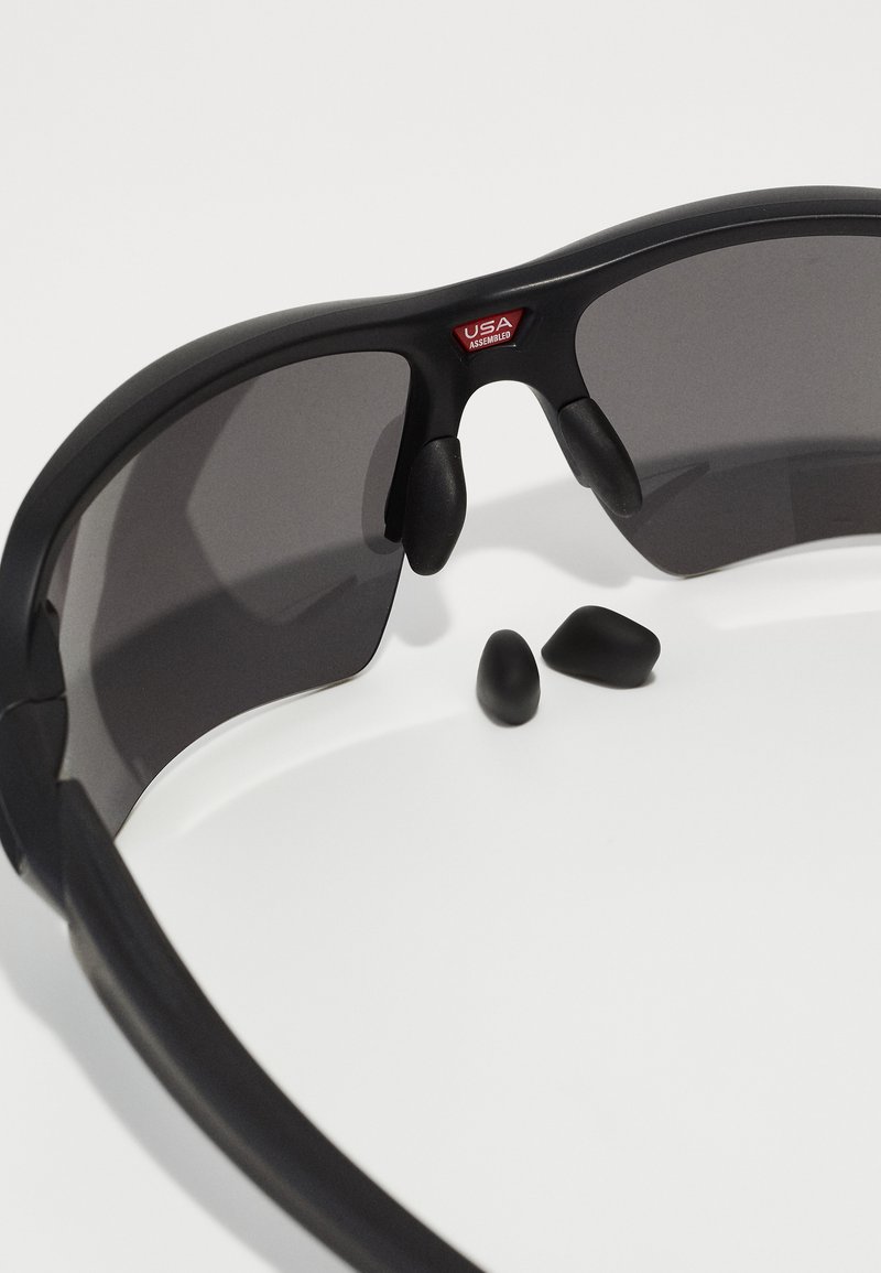 Black sunglasses with gray tinted lenses, featuring rubber nose pads and removable black ear tips, complementing the sleek, sporty design.