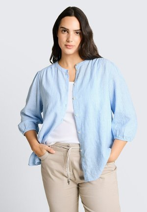 Frau mit langen dunklen Haaren, die ein hellblau gestreiftes Hemd mit Knöpfen über einem weißen Top und beigefarbenen Hosen trägt, die Hände in den Taschen.