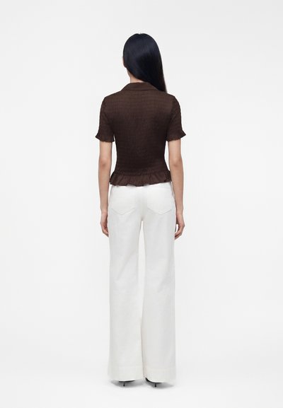 Femme aux longs cheveux noirs portant un haut marron cintré à manches courtes et un pantalon blanc taille haute à jambes larges, debout de dos.