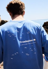 Modrá košile s dlouhým rukávem, na zadní straně s bílým potiskem ptáků a textem. Měkká tkanina s volným střihem a kulatým výstřihem.