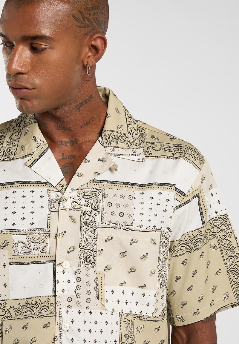 Chemise à manches courtes beige avec un design patchwork. Présente plusieurs motifs en noir et blanc, avec une fermeture boutonnée à l'avant et un col.