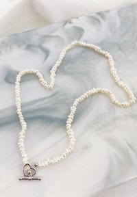 Witte parelketting met onregelmatig gevormde kralen, voorzien van een zilveren hartvormige sluiting. Minimalistisch ontwerp met een gladde textuur.