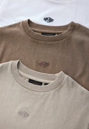 Drie gestapelde t-shirts met korte mouwen: één wit, één donkerbruin en één lichtbeige. Elk heeft een geribbelde halslijn en een geborduurd logo.