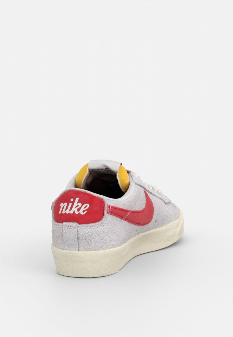 Sneaker in suede grigio con accenti rossi, caratterizzata da un logo ricamato bianco sul tallone e un interno giallo. Suola in gomma con dettagli testurizzati.