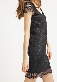 Kvinna som bär en figursydd svart spetsklänning med korta ärmar och vågig nederkant, som står framför en enfärgad vit bakgrund.