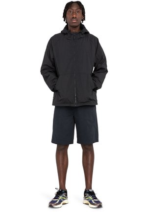 Black zip-up jacket with hood, two front pockets, paired with dark shorts and multi-colored sneakers. Lightweight material, casual fit.