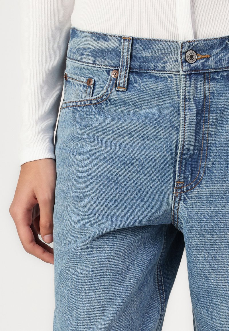 Hellblaue Denim-Jeans mit lockerer Passform, fünf Taschen, Kupferbeschlägen und hellen Nähdetails entlang des Taillenbands und der Taschen.