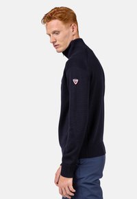Maglione di maglia blu navy con colletto alto e polsini a costine, con una toppa del logo sulla spalla sinistra. Materiale strutturato con design aderente.