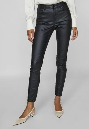Woman wearing black faux leather skinny pants, white long-sleeve blouse, and beige pointed slingback heels, standing against gray background.
