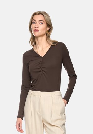 Frau mit blondem Haar, die ein braunes langärmeliges V-Ausschnitt-Oberteil und hellbeige, fein gestreifte Hose trägt, mit einer Hand in der Tasche.