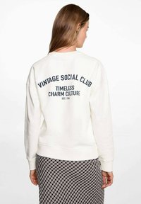 Woman with long brown hair wearing a white sweatshirt with "Vintage Social Club" text and a patterned skirt, facing away against a white background.