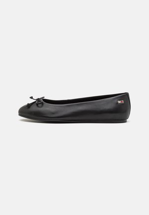 Ballerine plate en cuir noir avec un petit nœud sur l’orteil et un petit logo rectangulaire sur le côté du talon sur fond blanc.