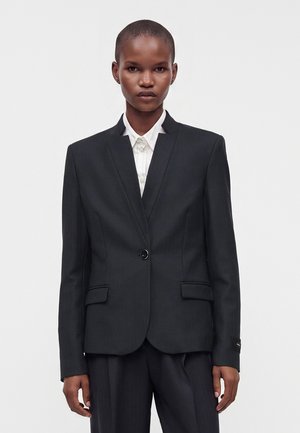 Kvinde med kort hår iført en sort enkeltknappet blazer og hvid skjorte med krave mod en ensfarvet baggrund.