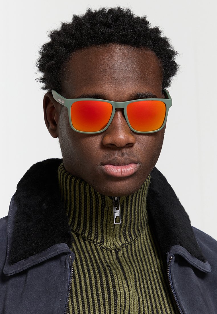 Zielone okulary przeciwsłoneczne z lustrzanymi czerwonymi soczewkami. Kwadratowy kształt oprawki o matowym wykończeniu. Noszone przez osobę w swetrze z wysokim kołnierzem.