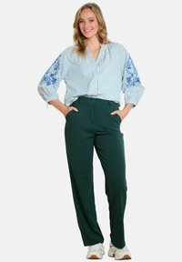 Camicia a righe abbottonata in azzurro chiaro e bianco con ricami floreali blu sulle maniche, abbinata a pantaloni aderenti verdi scuri e scarpe bianche.