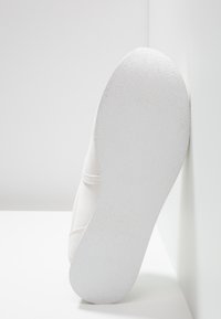 Baskets blanches posées la semelle vers le haut contre un mur blanc sur une surface blanche, montrant une semelle texturée et un design minimaliste.