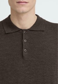 Sötétbarna polo póló, texturált kötött anyagból készült. Nyakgallérral és három barna gombbal rendelkezik a nyaknál.