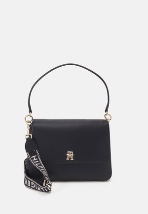 Sac à épaule en cuir noir avec emblème logo doré, poignée courte et bandoulière large en tissu détachable avec texte blanc.