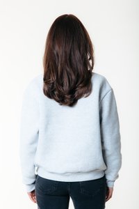 Persona con cabello largo castaño, vistiendo una sudadera gris claro y jeans oscuros, de pie con la espalda hacia la cámara contra un fondo blanco.