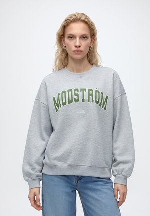 Femme aux cheveux blonds portant des boucles d'oreilles en or, un sweat-shirt gris avec un texte vert "MODSTRÖM" et un jean bleu, debout devant un fond uni.