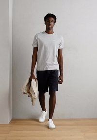 Tricou gri cu mânecă scurtă, pantaloni scurți negri și adidași albi. Ține o jachetă bej. Design simplu, material neted, culori neutre.