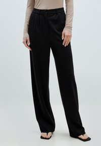 Zwarte wijde broek van zacht materiaal, met plooien, een hoge taille en een op maat gemaakte pasvorm. Gecombineerd met een beige lange mouw top en zwarte sandalen.