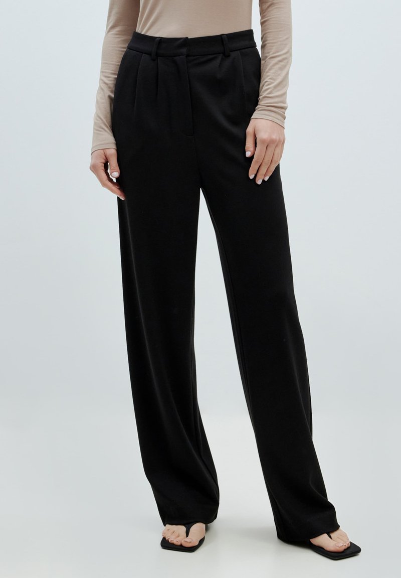 Zwarte wijde broek van zacht materiaal, met plooien, een hoge taille en een op maat gemaakte pasvorm. Gecombineerd met een beige lange mouw top en zwarte sandalen.