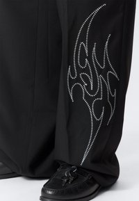 Μαύρο παντελόνι με ασημένια κεντήματα που θυμίζουν φλόγες στο πλάι, κατασκευασμένο από λείο ύφασμα. Συνδυασμένο με μαύρα μοκασίνια και κλειδιά που φαίνονται κοντά.