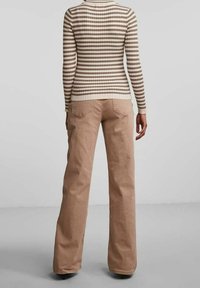 Osoba ubrana w beżowe spodnie o szerokich nogawkach oraz dopasowany sweter z długim rękawem w brązowo-białe paski, stojąca tyłem na jednolitym tle.
