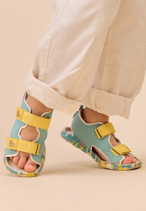 Türkisfarbene Sandalen mit gelben verstellbaren Riemen, offenem Zehendesign, gemusterter Sohle mit blauen, gelben und grünen Akzenten, aus weichem Material gefertigt.