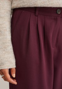 Ruka odmara blizu bordo visokih plišanih hlača s dugmetom i petljama za pojas, u bež pletenom džemperu i srebrnom prstenom.