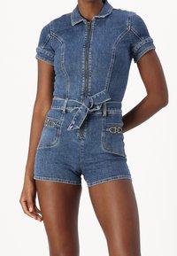 Kvinna som bär en blårandig denimshorts med kort ärm, dragkedja framtill, knytbälte och två framfickor med metallic spänndetaljer.