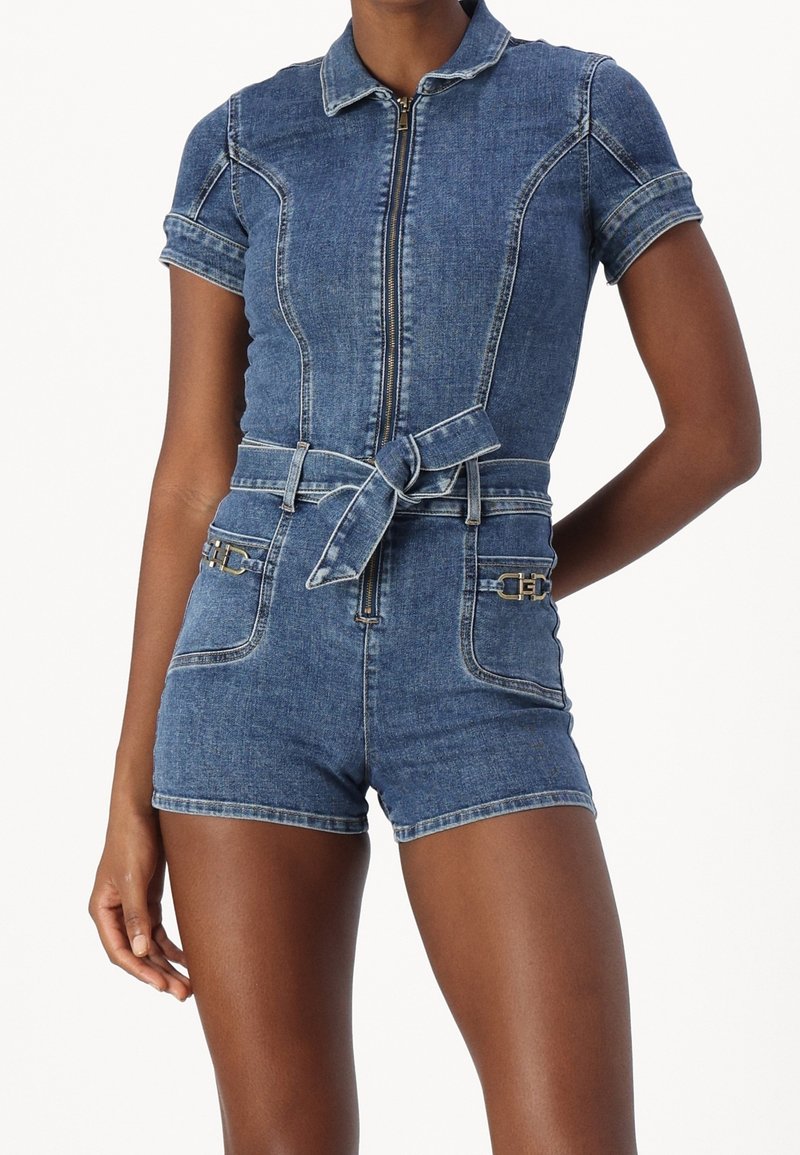 Kvinna som bär en blårandig denimshorts med kort ärm, dragkedja framtill, knytbälte och två framfickor med metallic spänndetaljer.