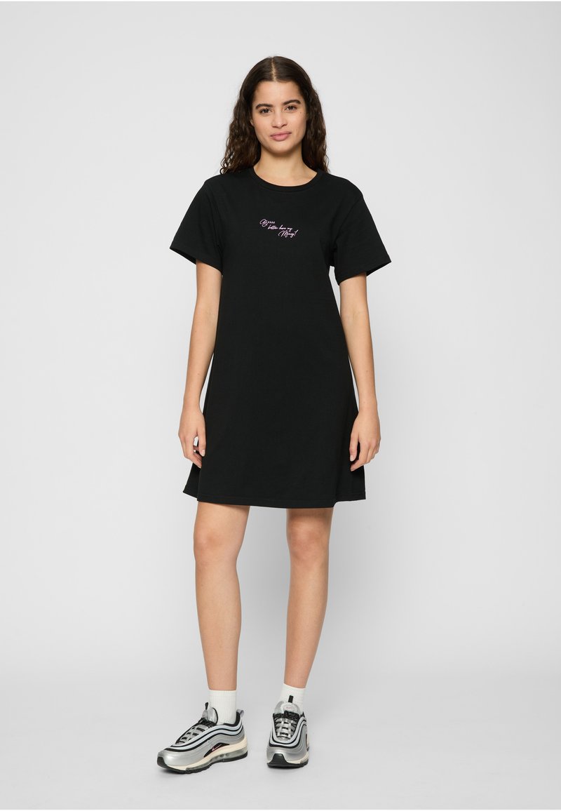 Femme debout dans une robe noire à manches courtes avec un texte écrit sur la poitrine et portant des chaussettes blanches avec des baskets argentées et noires.