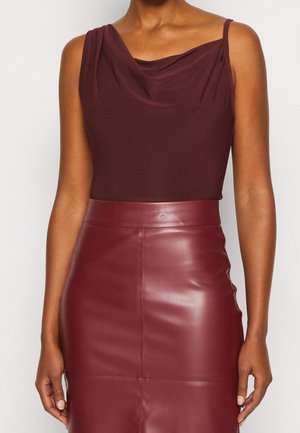 Vrouw draagt mouwloze bordeauxrode top met gedrapeerde hals, ingestopt in een hoog getailleerde bordeauxrode leren rok, staand tegen een witte achtergrond.