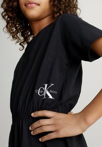 Musta puuvillamekko, jossa vyötärö kavennettu ja lyhyet hihat. Edessä näkyy valkoinen logo, jossa lukee "Calvin Klein Jeans".