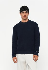 Marine gebreide trui met een gestructureerd oppervlak, ronde halslijn en geribbelde manchetten. Heeft een logo-details op de borst. Gekocht met lichtgekleurde broek.