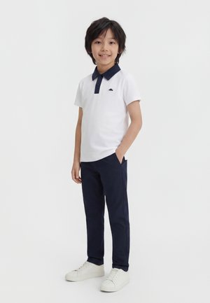 Boy standing and smiling, wearing a white polo shirt with navy collar, navy pants, and white sneakers on a plain white background.