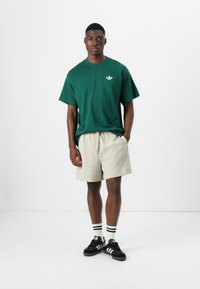 Un tricou verde din bumbac cu logo alb, pantaloni scurți bej cu model și adidași negri cu dungi albe, completați cu șosete negre și albe.