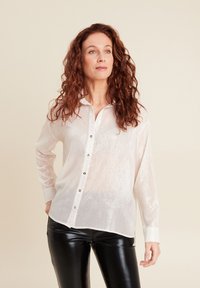 Camicia bianca trasparente con colletto, maniche lunghe e bottoni argentati. Abbinata a pantaloni in pelle nera attillati. Texture liscia.