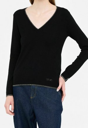 Femme portant un pull noir à col en V et manches longues avec bordure métallique, associée à un jean bleu en denim, main posée sur la hanche.
