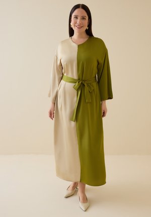 Donna sorridente che indossa un lungo vestito bicolore diviso beige e verde oliva con una cintura annodata abbinata e scarpe crema a punta.
